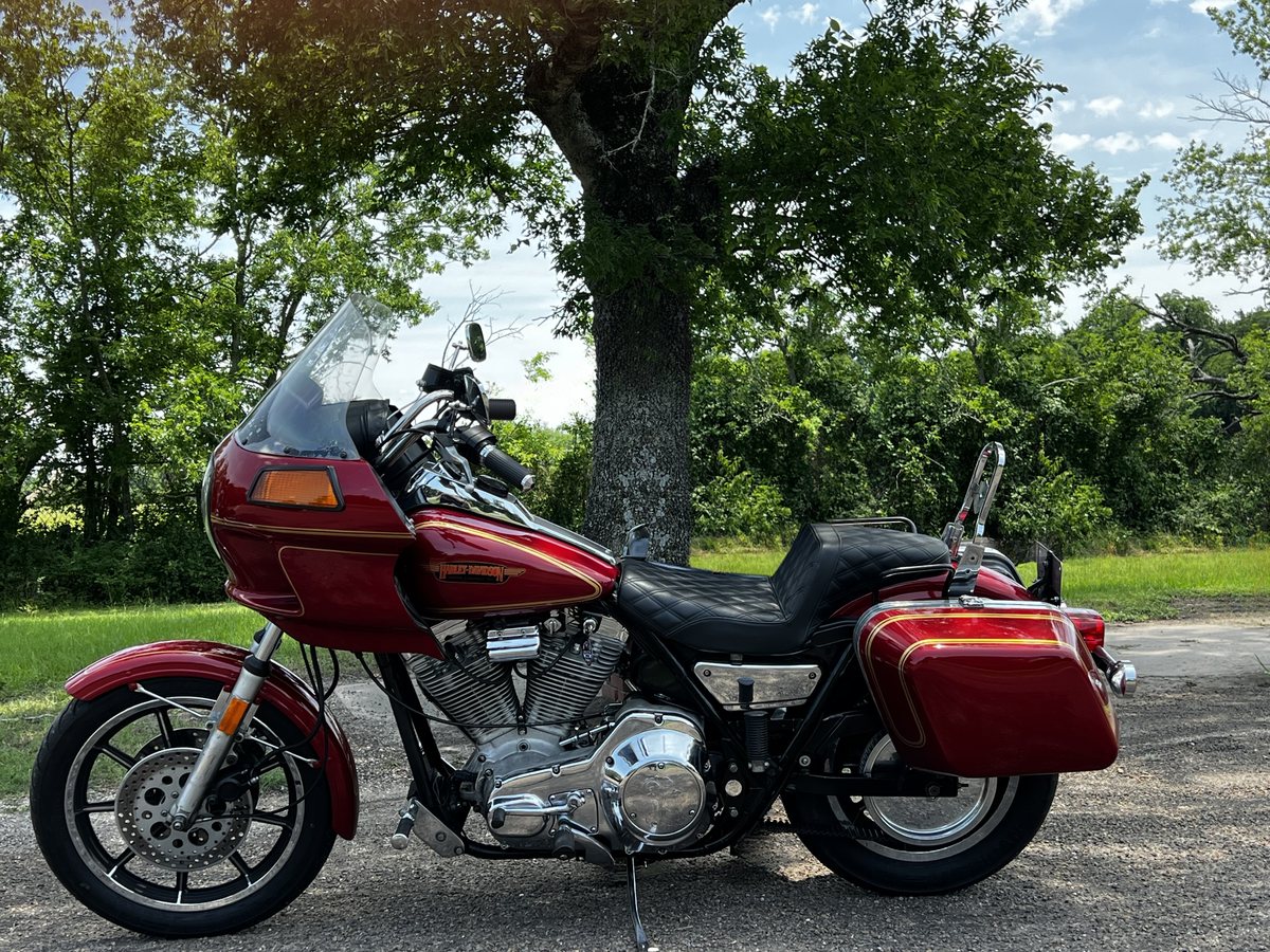 A red Harley-Davidson FXRT with fairing and hard saddlebags, seen from the left side, parked on gravel under a large tree