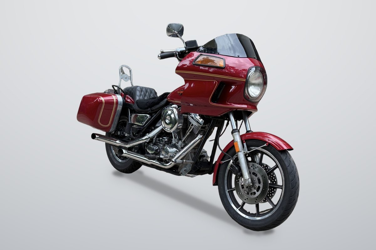 Front three-quarter view of a red Harley-Davidson FXRT with a gold-striped fairing, round headlight and chrome exhaust, on a plain grey backdrop