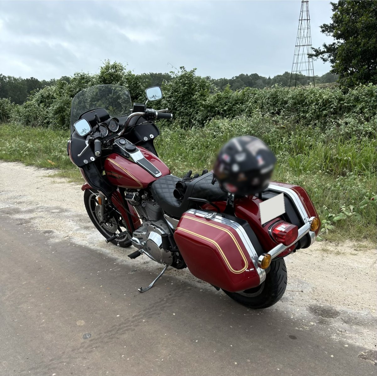 A red Harley-Davidson FXRT parked on the shoulder of a country road, seen from behind, with gold-striped saddlebags and a short windscreen